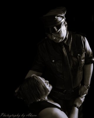 This is a black and white photo. Master SirTee is dressed in a black leather shirt, tie, and pants, and is wearing eyeglasses and a Master's cover. He is standing looking down at little kitsune. little kitsune's face is turned up looking back at Him. Master SirTee has one hand on the side of little kitsune's head and the other is holding her hand. little kitsune is wearing a sleeveless black shirt, has medium length bobbed hair.