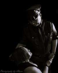 This is a black and white photo. Master SirTee is dressed in a black leather shirt, tie, and pants, and is wearing eyeglasses and a Master's cover. He is standing looking down at little kitsune. little kitsune's face is turned up looking back at Him. Master SirTee has one hand on the side of little kitsune's head and the other is holding her hand. little kitsune is wearing a sleeveless black shirt, has medium length bobbed hair.